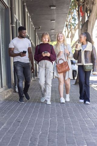Diverse friends walking together on city sidewalk holding coffee and using smartphones