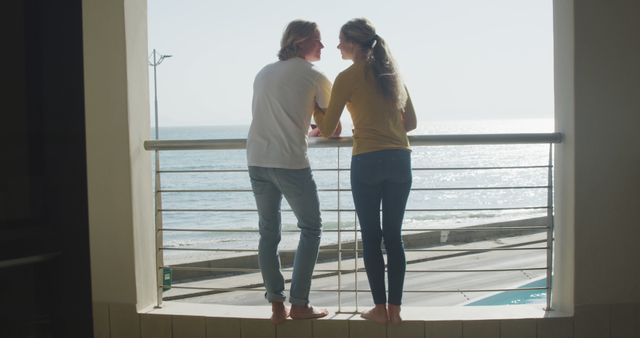 Young couple standing on a balcony, overlooking the ocean, enjoying the serene view. They are wearing casual clothing, appearing relaxed and content. Can be used for themes of couple's retreats, romantic getaways, relaxation, leisure time, or seaside vacations.