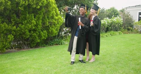 Graduates celebrating on campus lawn taking selfie in caps and gowns holding diplomas