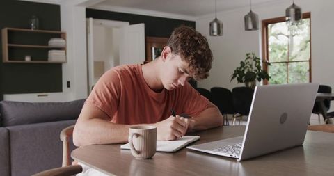 Young man writing at dining table with laptop and coffee, planning and focusing at home
