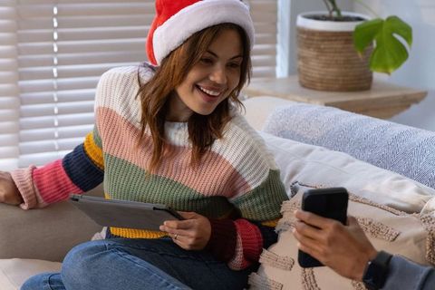 Couple celebrating christmas at home woman in santa hat holding tablet partner using phone
