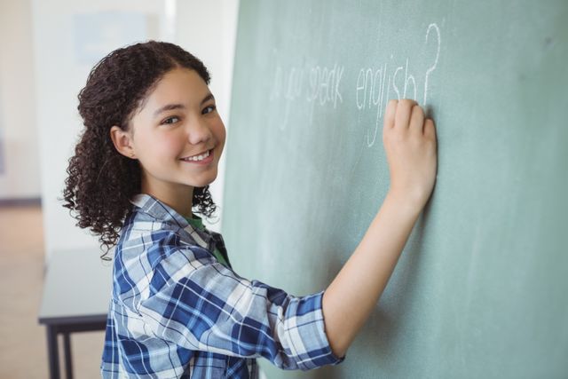 Young schoolgirl with curly hair smiling while writing on a chalkboard in a classroom. Ideal for educational materials, school promotions, and articles about learning and teaching. Can be used to depict student engagement, classroom activities, and academic environments.