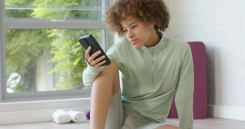African american woman checking smartphone while sitting by sunlit window after workout