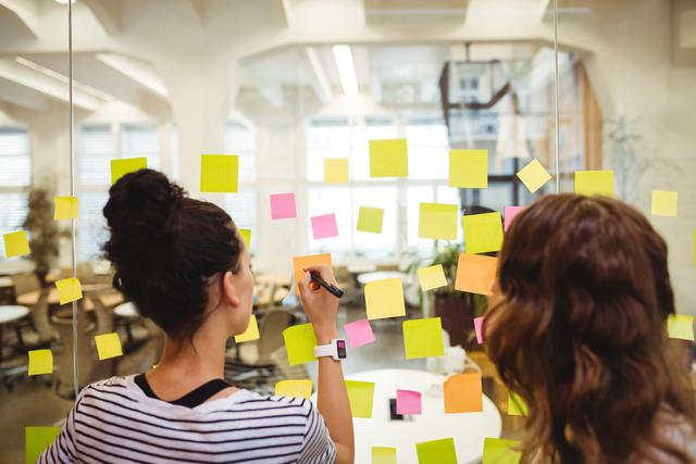 Business executives engaging in a brainstorming session using colorful sticky notes on a glass wall in a modern office. Ideal for illustrating concepts of teamwork, collaboration, planning, and creative strategy sessions in corporate environments.