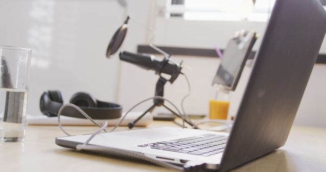 Modern workspace featuring a laptop, microphone, headphones, and glass of water. Ideal for illustrating concepts such as remote work, online meetings, podcast creation, and home office setups.