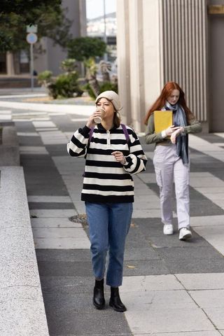 Two women walking on urban sidewalk drinking coffee while carrying backpacks