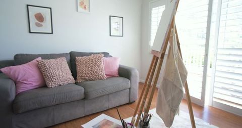 Sunlit creative living room featuring gray sofa, pink cushions and artist easel