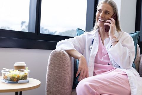Female clinician taking break in clinic lounge in lab coat with stethoscope, smiling on phone