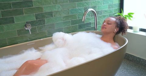 Woman relaxing in freestanding bathtub with thick bubbles and green tile spa vibe
