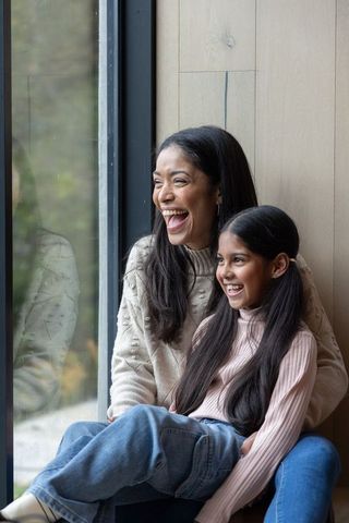 Asian mom and daughter laughing on window seat, cozy sweaters, natural light, family warmth