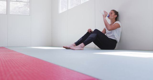 Woman sitting against wall in gym resting after workout, drinking water to stay hydrated. Useful for fitness and wellness articles, gym membership promotions, and exercise-related websites.