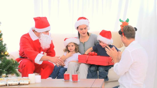 Santa Claus is sitting in a family living room, offering a gift to a young boy, creating a joyful and festive moment. The boy is surrounded by family members, including his parents and possibly grandparents, all dressed in holiday attire with Santa hats, sitting by a decorated Christmas tree. This could be used for promotional materials, holiday greeting cards, or content related to family celebrations and Christmas traditions.