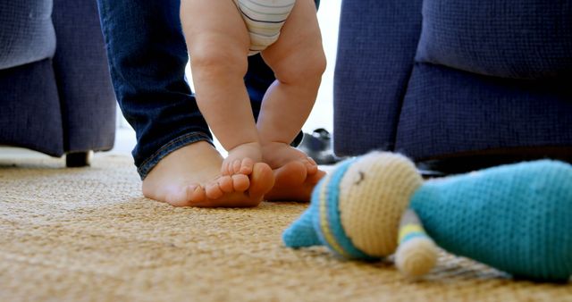 A young child takes tentative steps while holding onto an adult's legs, with a plush toy lying on the carpet nearby. It captures a milestone moment of a baby's first steps, symbolizing growth and parental support.