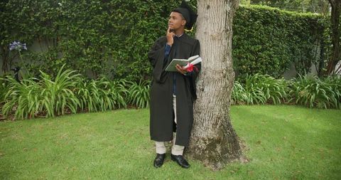 Graduating Student Leaning on Tree Holding Diploma and Reflecting in Lush Garden Lawn