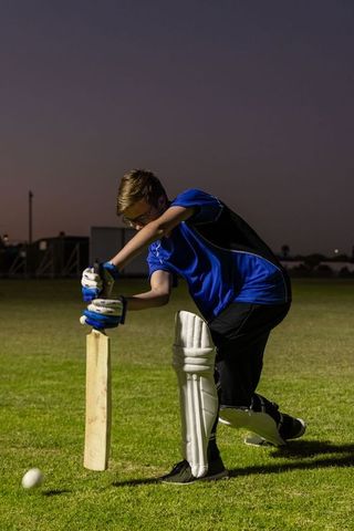 Teen cricketer defending during twilight practice on grass pitch with bat and pads