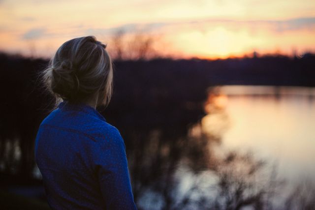 Woman standing by river, watching sunset. Ideal for themes like peace, contemplation, nature, and quiet moments. Perfect for use in blogs, advertisements, posters, and social media promoting outdoor activities, mindfulness, and relaxation.