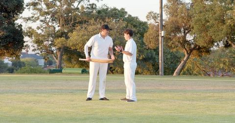Two cricket players discussing strategy at sunset on grassy park field wearing whites