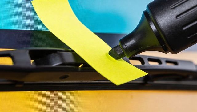 Close-up chisel-tip marker pressing yellow sticky note on windshield wiper with teal warm bokeh