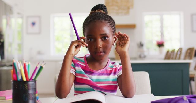 A young African American girl is sitting at a table, holding a pencil and pondering while doing her homework. She is surrounded by colorful pencils and books, indicating an educational or study environment. This image can be used for educational websites, tutoring services, school-related promotional materials, and articles focusing on children's learning and development at home.