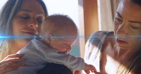 Sunlit morning bonding between two mothers cradling newborn with gentle lens flare