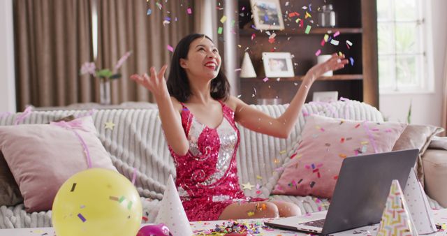 Image of smiling biracial woman throwing confetti dressed in pink dress. Party, leisure time, domestic life and lifestyle concept.