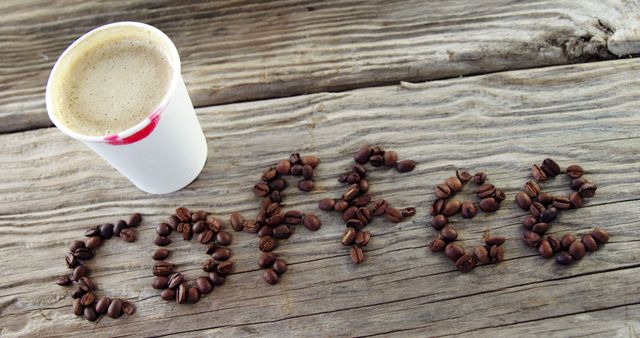 Coffee beans are arranged on a rustic wooden surface to spell the word COFFEE, next to a paper cup filled with a frothy beverage, with copy space. The arrangement conveys a creative and inviting message for coffee lovers and highlights the beverage's popularity.