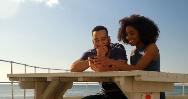African American couple sitting on wooden bench by the beach, sharing a laugh while looking at a smartphone. Sun shining brightly, creating a relaxed summer vibe. Perfect for content related to travel, leisure, relationships, and technology. Could be used for advertisements promoting vacation destinations, outdoor activities, or digital devices.
