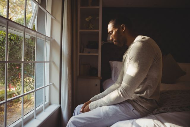 Man sitting on bed by window, looking thoughtful and sad. Sunlight streaming through window, creating a serene yet melancholic atmosphere. Ideal for use in articles or campaigns related to mental health, depression, loneliness, or personal reflection.