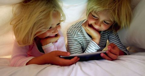 Happy Siblings Using Tablet Under Blanket Relax in Cozy Bedroom