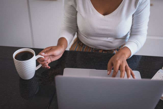 This image depicts a biracial woman working on a laptop while holding a cup of coffee at home. Ideal for articles or blogs about remote work, productivity, quarantine lifestyle, and modern home office setups. Can also be used in advertisements for coffee, technology, or home office furniture.