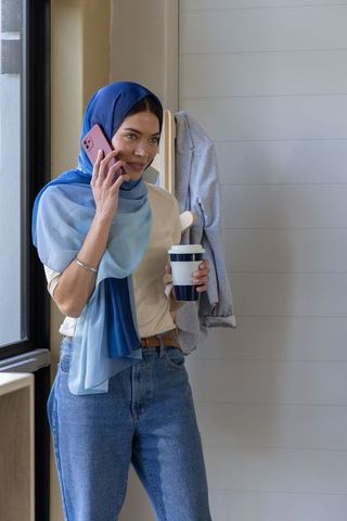 Modern woman wearing blue hijab talking on phone holding travel mug in entryway