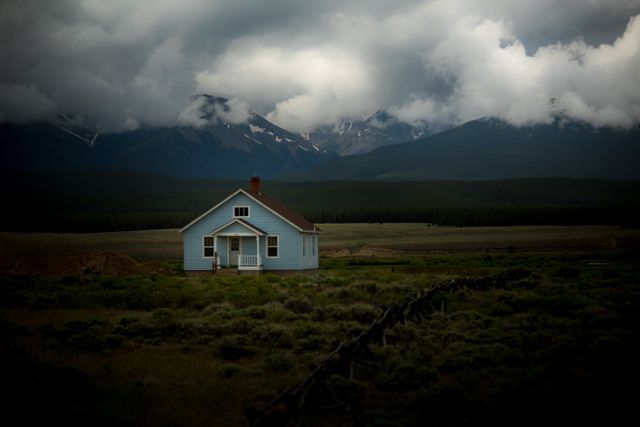 Charming blue house located in a scenic landscape with mountains in the background. Storm clouds create a dramatic sky, contrasting the serene green fields around the house. Suitable for themes of isolation, tranquility, rural living, nature's beauty, and storm watching.