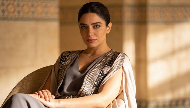 Regal woman wearing embroidered ceremonial robe seated on upholstered chair in warm sunlit interior