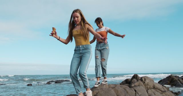 Two teenage girls exploring rocks at the beach while enjoying ice cream. Both are wearing casual clothing, having fun, and balancing on the rocks. Perfect for themes related to friendship, summertime activities, and outdoor adventures. Ideal for marketing campaigns, travel blogs, and lifestyle articles.