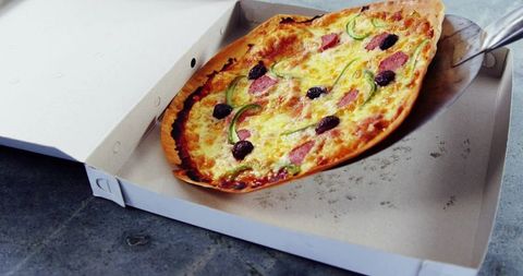 Serving thin-crust pizza with pepperoni and olives on metal spatula above takeout box