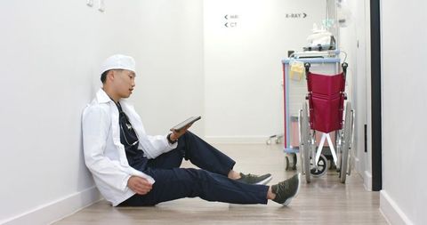 Asian doctor sitting in hospital corridor using tablet wearing white coat and stethoscope