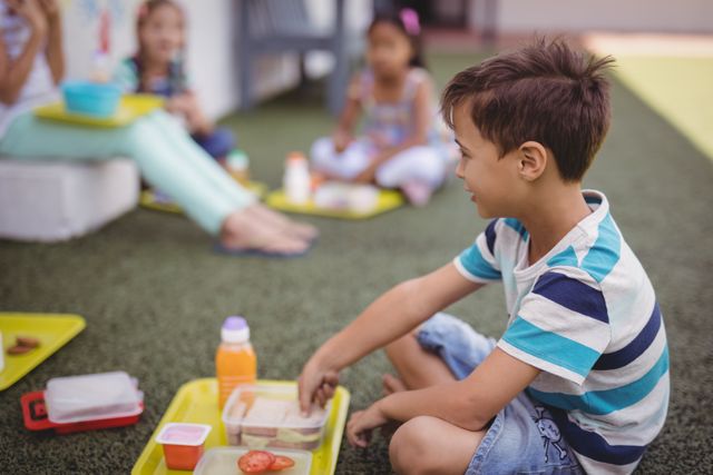 Schoolboy enjoying lunch outdoors with friends on a playground. Ideal for educational content, school lunch programs, healthy eating campaigns, and social interaction themes.