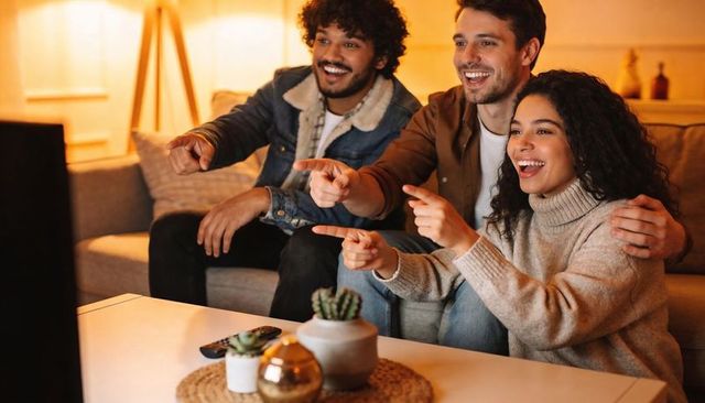 Friends laughing and pointing at tv while watching movie in cozy warm living room