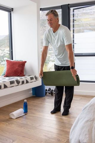 Mature man unrolling green yoga mat in sunlit bedroom for home workout and mobility