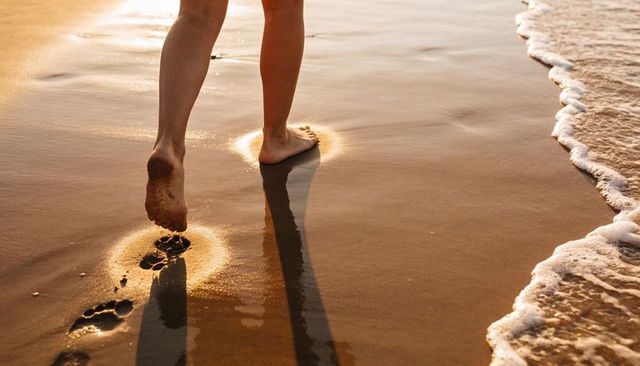 Sunlit barefoot walking along wet sand leaving footprints on golden shoreline