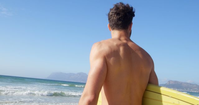A young Caucasian man stands on the beach holding a surfboard, with copy space. His gaze towards the ocean suggests he is preparing for or reflecting on a surfing session.