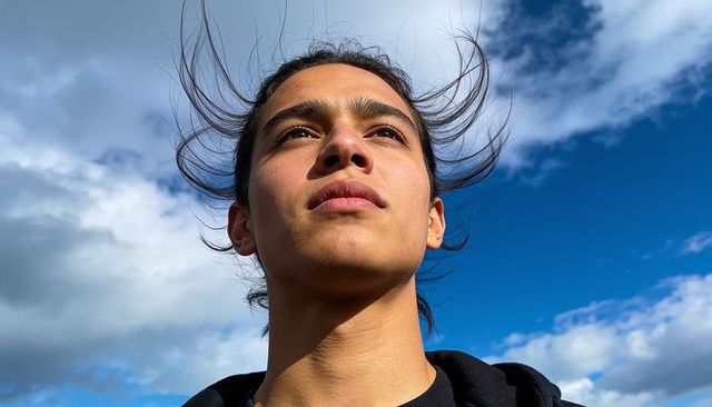 Young man gazing upward under windy blue sky with dramatic clouds, sunlit portrait