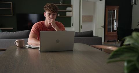 Young man working from home at laptop on dining table with coffee and notebook