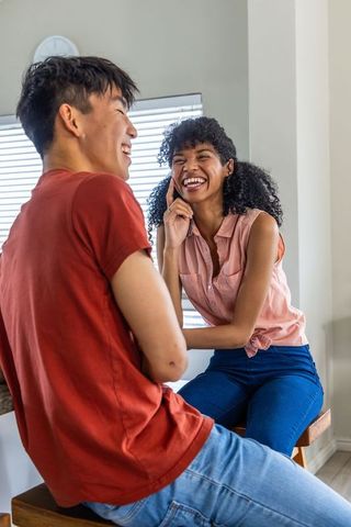 Multiracial couple laughing on stools sharing lighthearted conversation in sunlit home