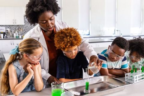 Teacher guiding children pouring green liquid into graduated cylinder during hands-on science lab