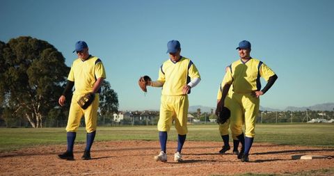 Senior baseball players walking infield wearing yellow jerseys and blue caps in sunlit park