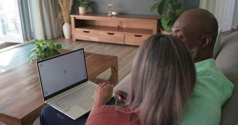 Diverse senior couple using laptop on couch in modern living room, staying connected