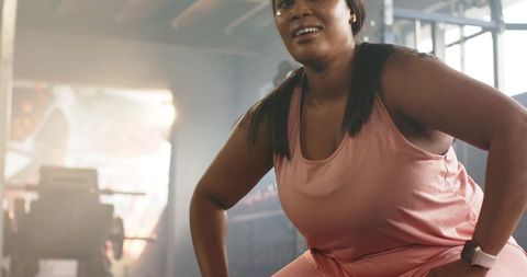 African american woman performing squats in backlit industrial gym wearing pink activewear