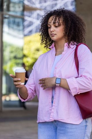 African american woman holding coffee by glass facade wearing pink shirt and smartwatch