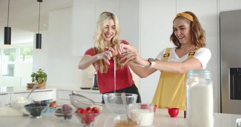 Two friends cracking egg into glass mixing bowl on sunlit kitchen island, baking together